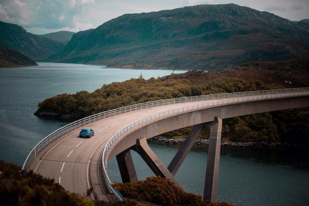 Un'auto blu attraversa un ponte curvo in un paesaggio di montagne e acqua
